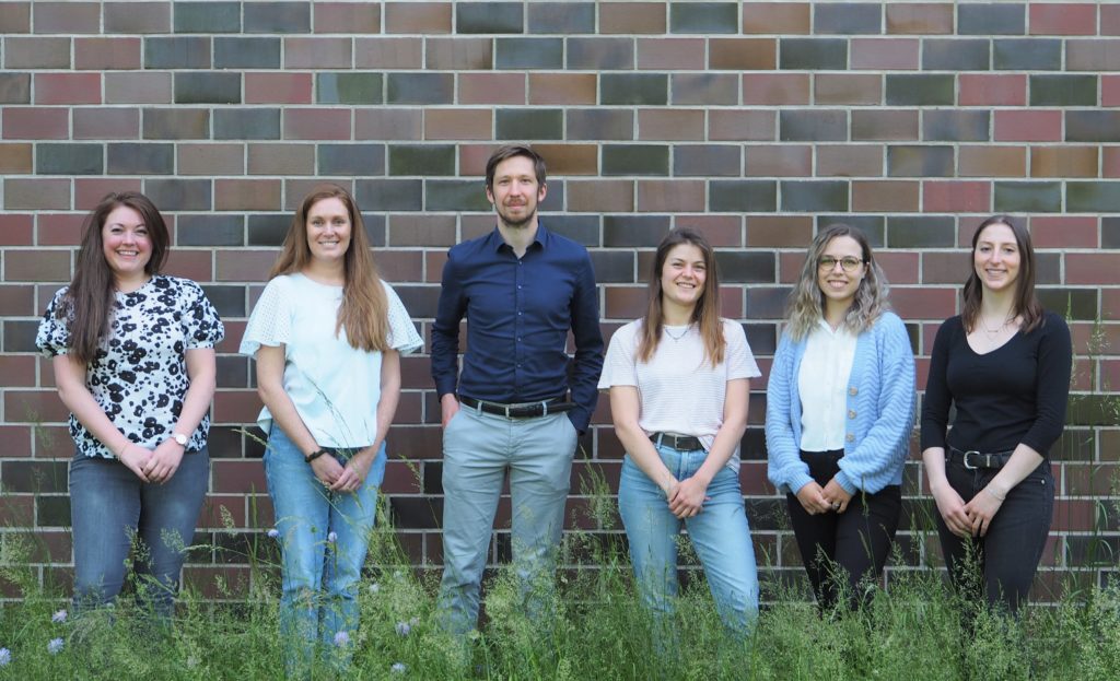 Six people standing against a cinderblock wall, facing the camera.