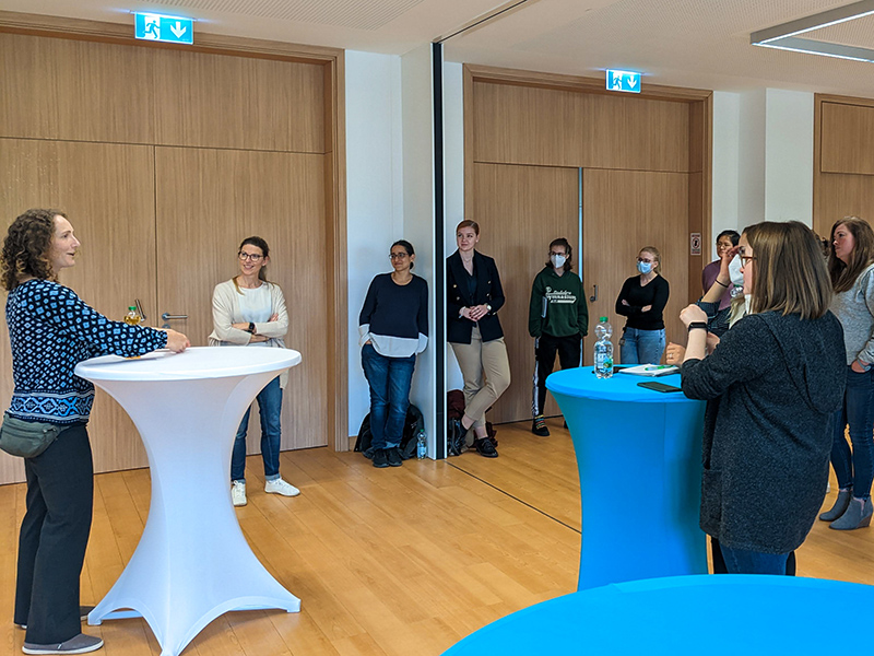 The photo shows a seminar room in the Chemikum. Various high tables with white and blue covers are set up, at which female students are standing and listening with interest. In the foreground, Prof. Young is talking about her academic career. All are casually dressed in jeans, sweaters, etc., a few of the female students are wearing mouth-nose protection and have beverage bottles in front of them.