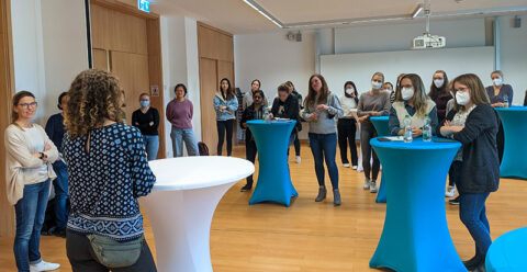 The photo shows a seminar room in the Chemikum. Various high tables with white and blue covers are set up, at which female students are standing and listening with interest. In the foreground, Prof. Young stands with her back to the photographer and talks about her academic career. All are casually dressed in jeans, sweaters, etc., a few of the female students are wearing mouth-nose protection and have bottles of drink in front of them.