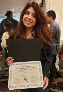 The photo shows Ms. Alkhashrom at the conference. She is smiling at the camera and holding up her certificate. The PhD student has long brown hair and is wearing a dark jacket.