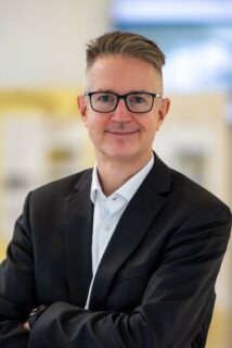 Portrait photo of Prof. Dr. Jörg Libuda. Mr. Libuda has short dark hair, shaved at the sides. He wears dark glasses, a black jacket and a white shirt. He smiles at the camera with his arms crossed in front of his chest. The light background of the photo is blurred.